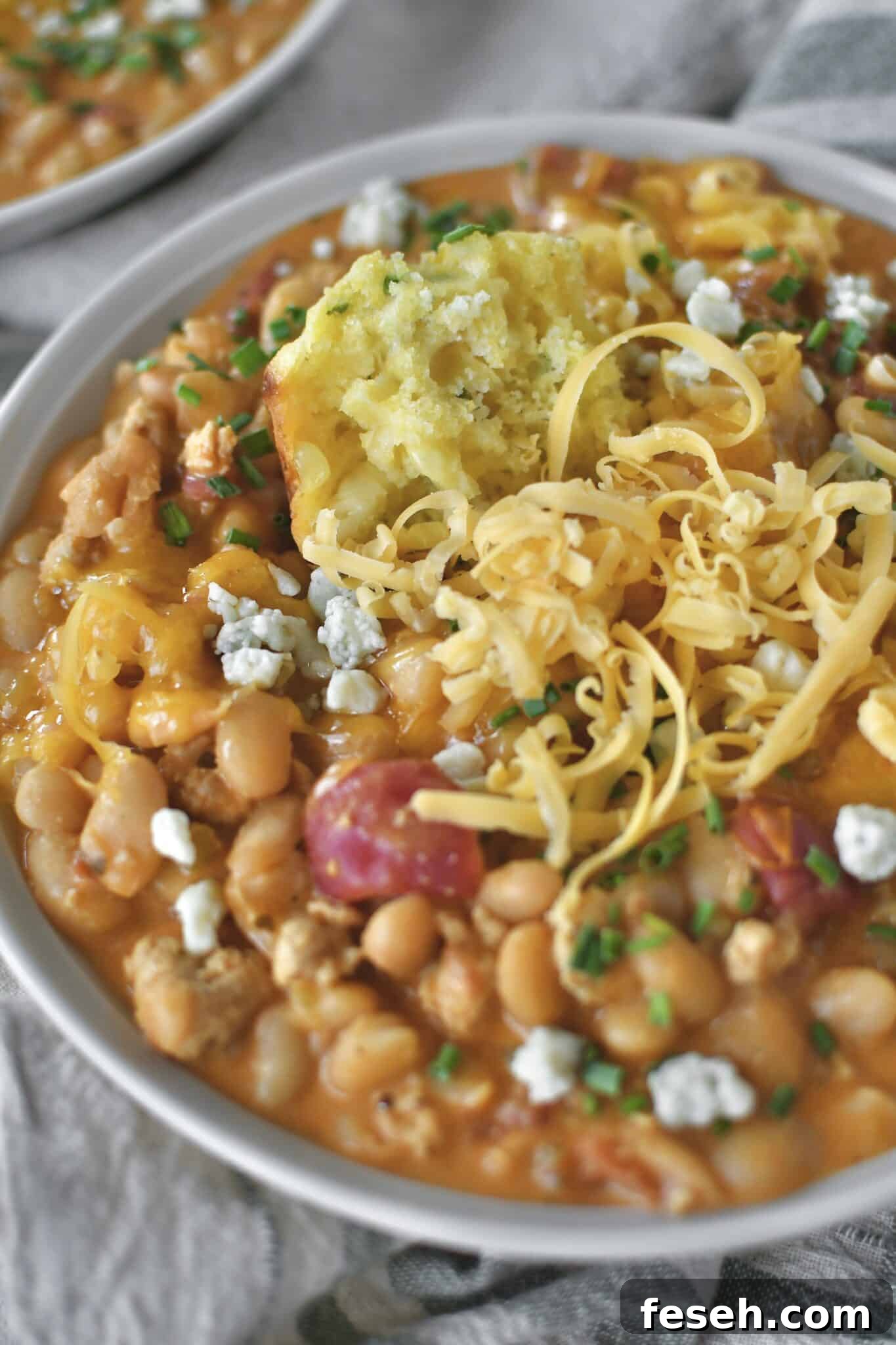Buffalo Chicken Chili served in a bowl, topped with chives, blue cheese, and cheddar cheese. And a piece of cornbread muffin dunked in it.