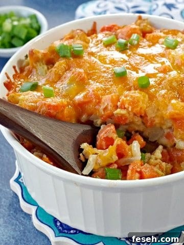 carrot casserole in white casserole dish with a brown wooden spoon scooping casserole (on blue background) with dish of green onions and oven mitts in the background