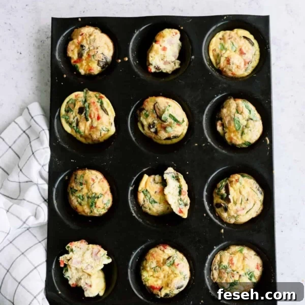 Muffin tray with twelve cooked egg muffins, some with spinach and vegetables, on a light surface next to a checkered cloth.