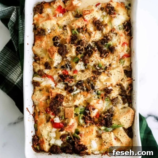 Baked casserole with bread cubes, bell peppers, onions, melted cheese, and browned sausage in a white rectangular dish on a green checkered cloth.
