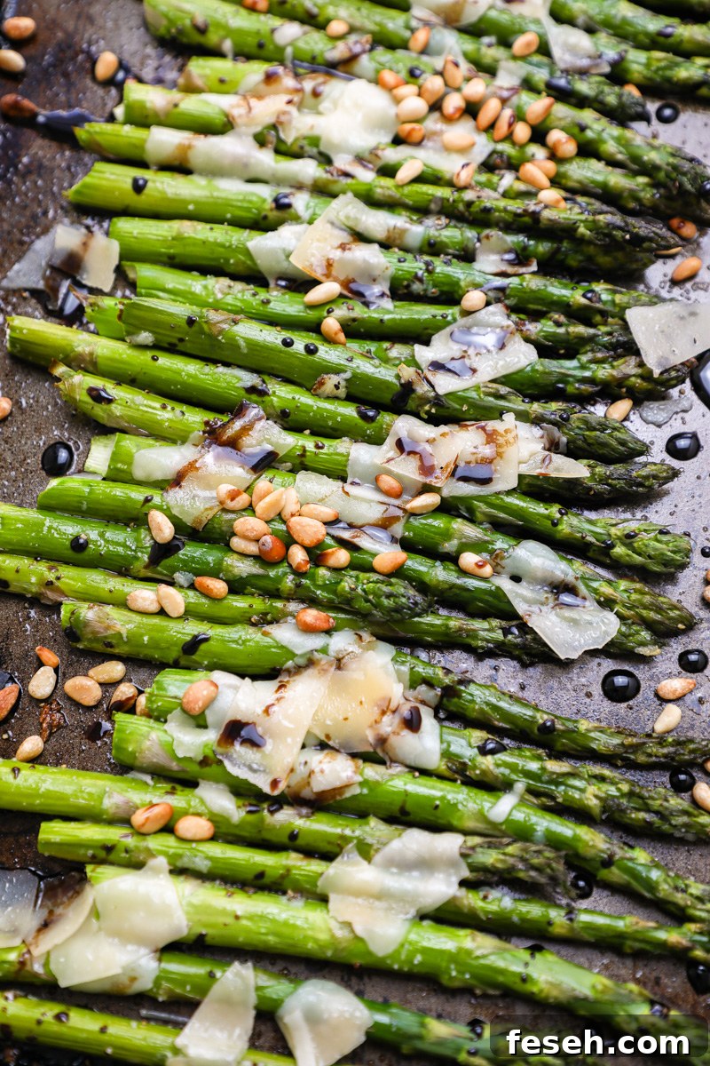 A dark aking pan filled with roasted asparagus topped with shaved parmesan, toasted pine nuts and balsamic glaze