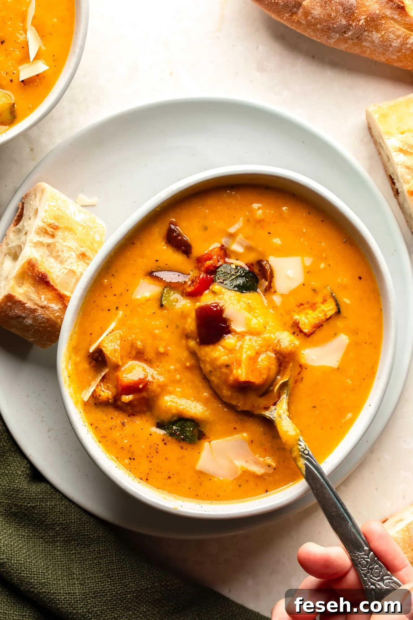 overhead view of a womans hand lifting a spoon out of a bowl of roasted vegetable soup.