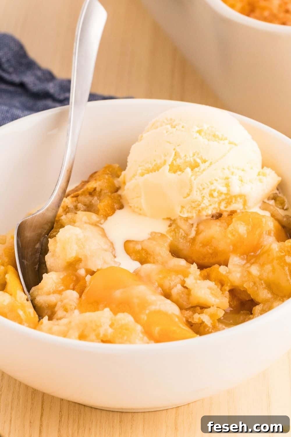 cake mix peach cobbler dump cake in a white bowl, topped with a scoop of vanilla ice cream. A spoon is in the bowl.