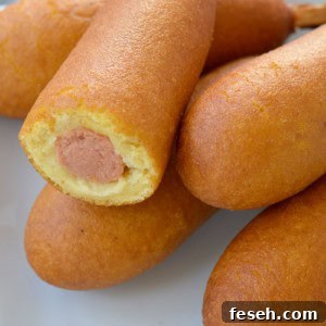 a pile of corn dogs on a white plate; one has a bite taken out