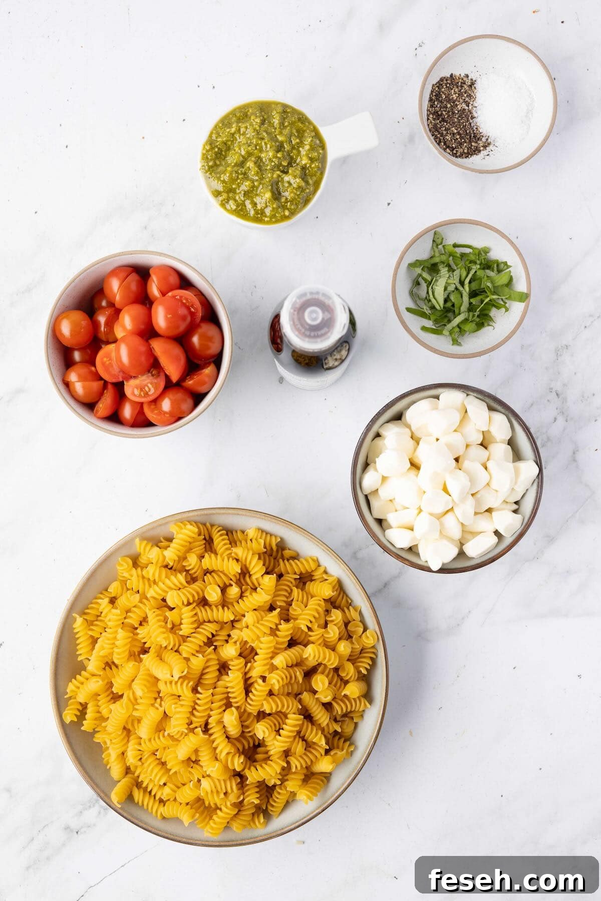 Ingredients for Caprese pasta salad including cherry tomatoes, mozzarella pearls, pesto, basil, and rotini pasta arranged on a kitchen counter.