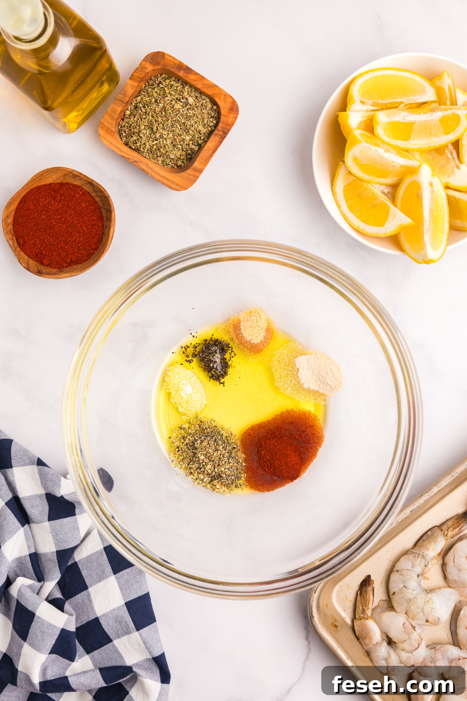 A glass mixing bowl containing olive oil and a variety of spices including paprika, oregano, onion powder, garlic powder, salt, and pepper. Ingredients are arranged around the bowl including lemon wedges, olive oil, and raw shrimp on a tray.