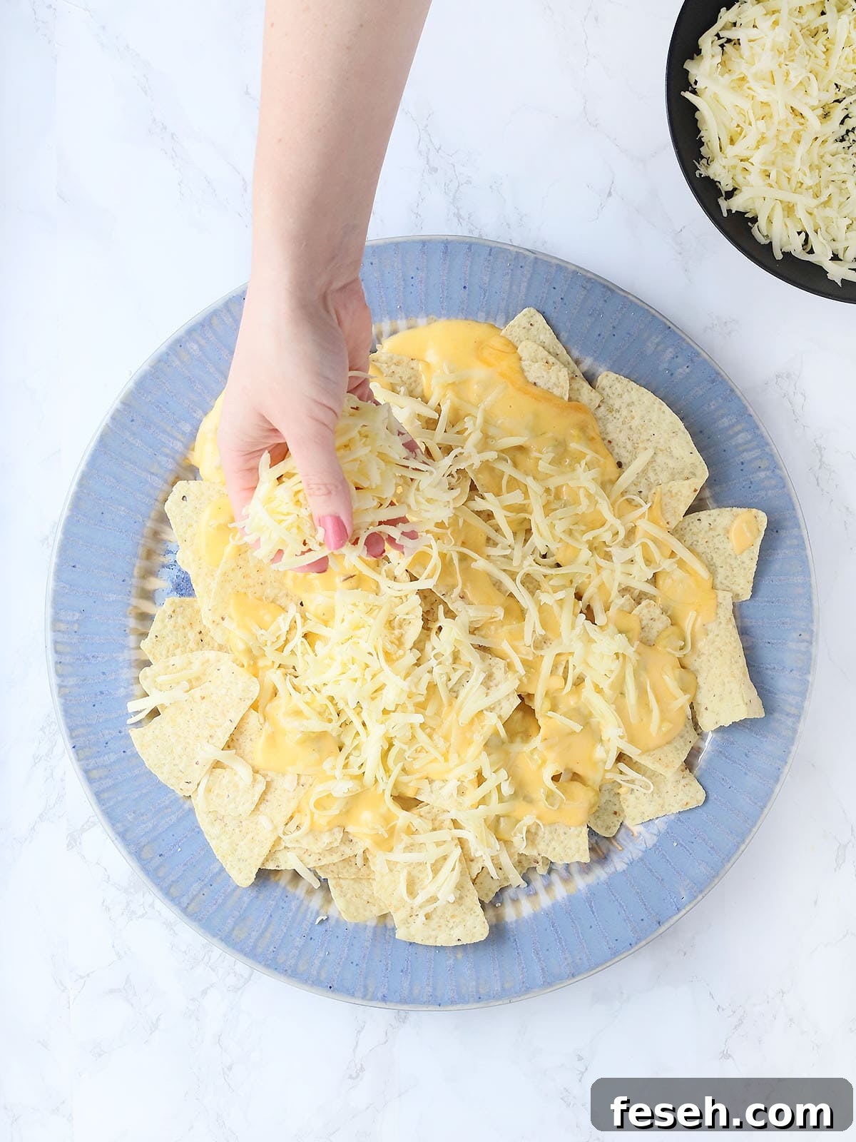 hand layering shredded Monterrey Jack cheese over a blue platter topped with tortilla chips and chili cheese sauce