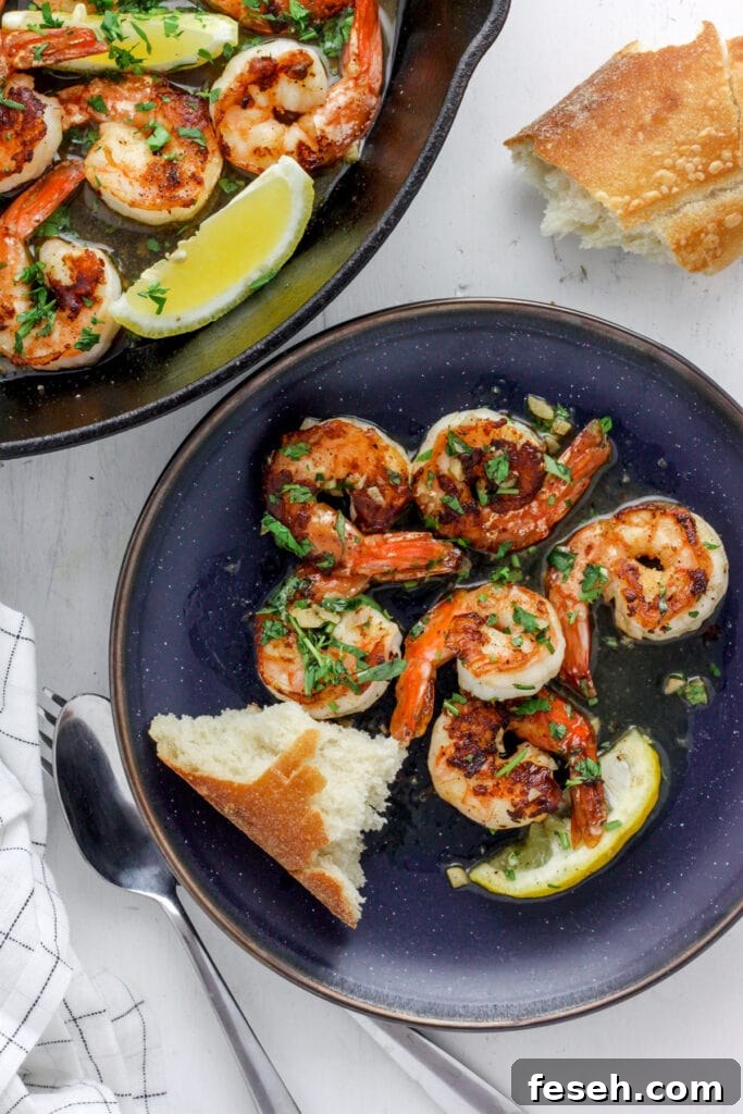 A blue plate with seared shrimp and crusty bread. off center is a skillet with more seared shrimp.