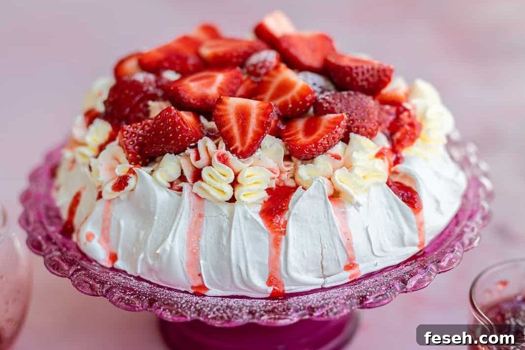 A meringue cake pavlova topped with whipped cream and fresh strawberry slices, drizzled with strawberry sauce, sits on a pink glass cake stand.