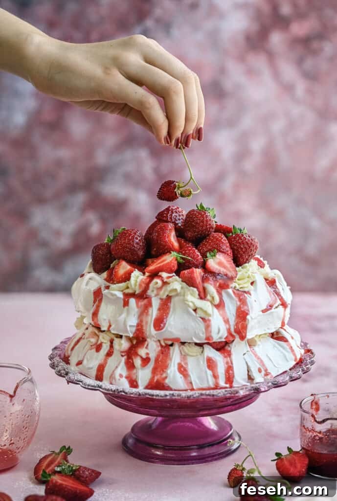 A hand places a fresh strawberry atop a two-layer meringue cake covered with whipped cream, strawberry sauce, and whole strawberries, on a pink glass cake stand.
