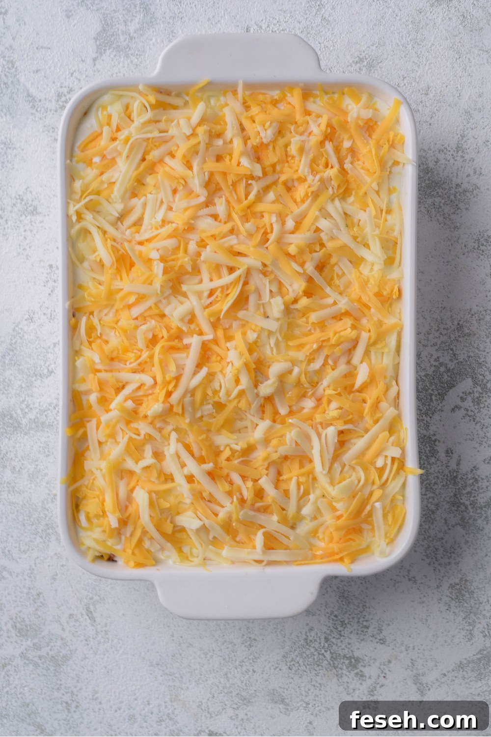 A white casserole dish with layered cornbread, beef, and shredded cheese before baking