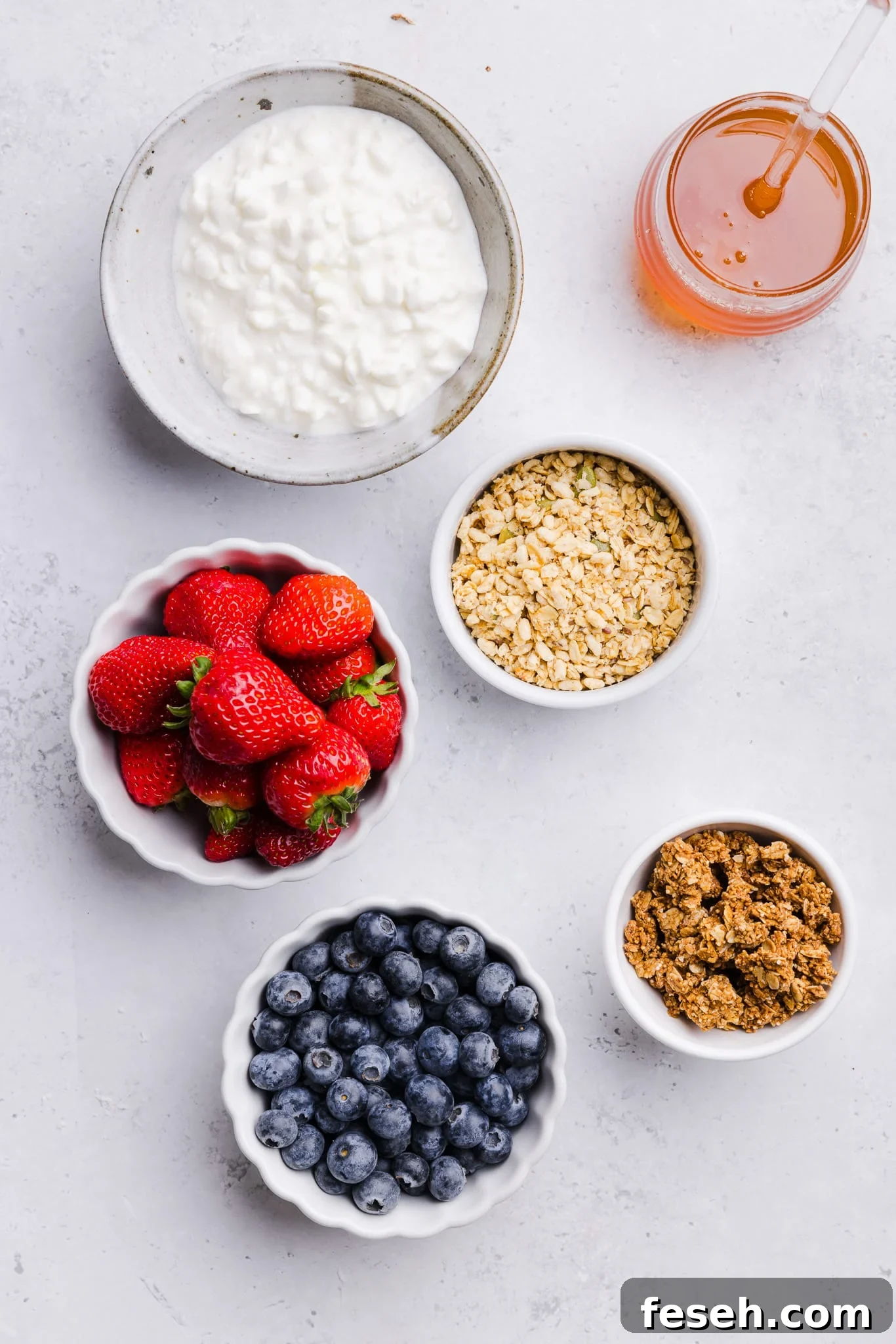 Cottage cheese, strawberries, blueberries, honey and granola in small bowls on a countertop.