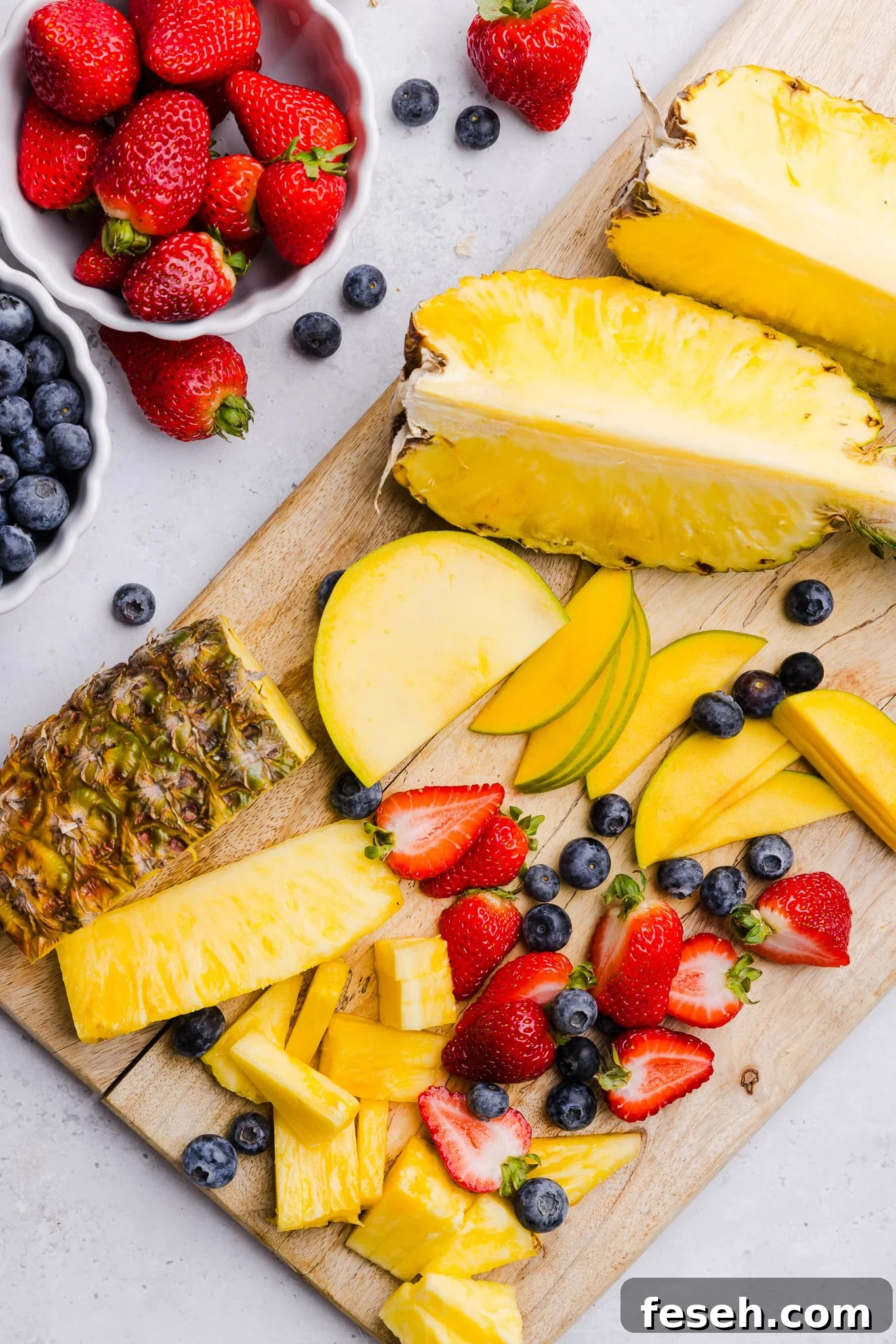 Pineapple, mango, strawberries and blueberries being sliced on a wooden cutting board for cottage cheese fruit bowls.