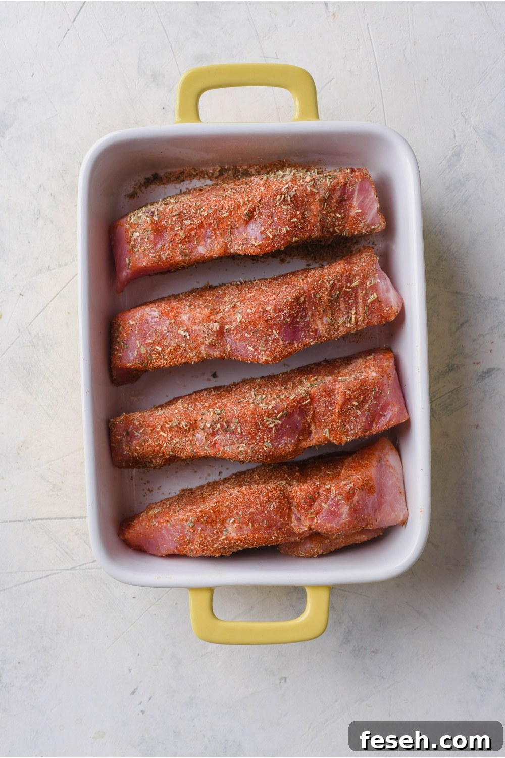Overhead view of four seasoned and boneless ribs placed evenly in a white baking dish with yellow handles.