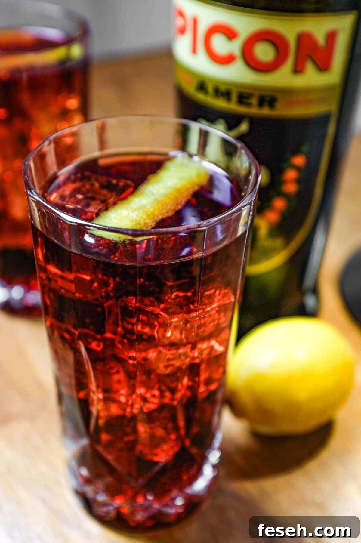 Two cut glass highball glasses of a cocktail called Picon Punch sit alongside a bottle of Picon Amer and a lemon.