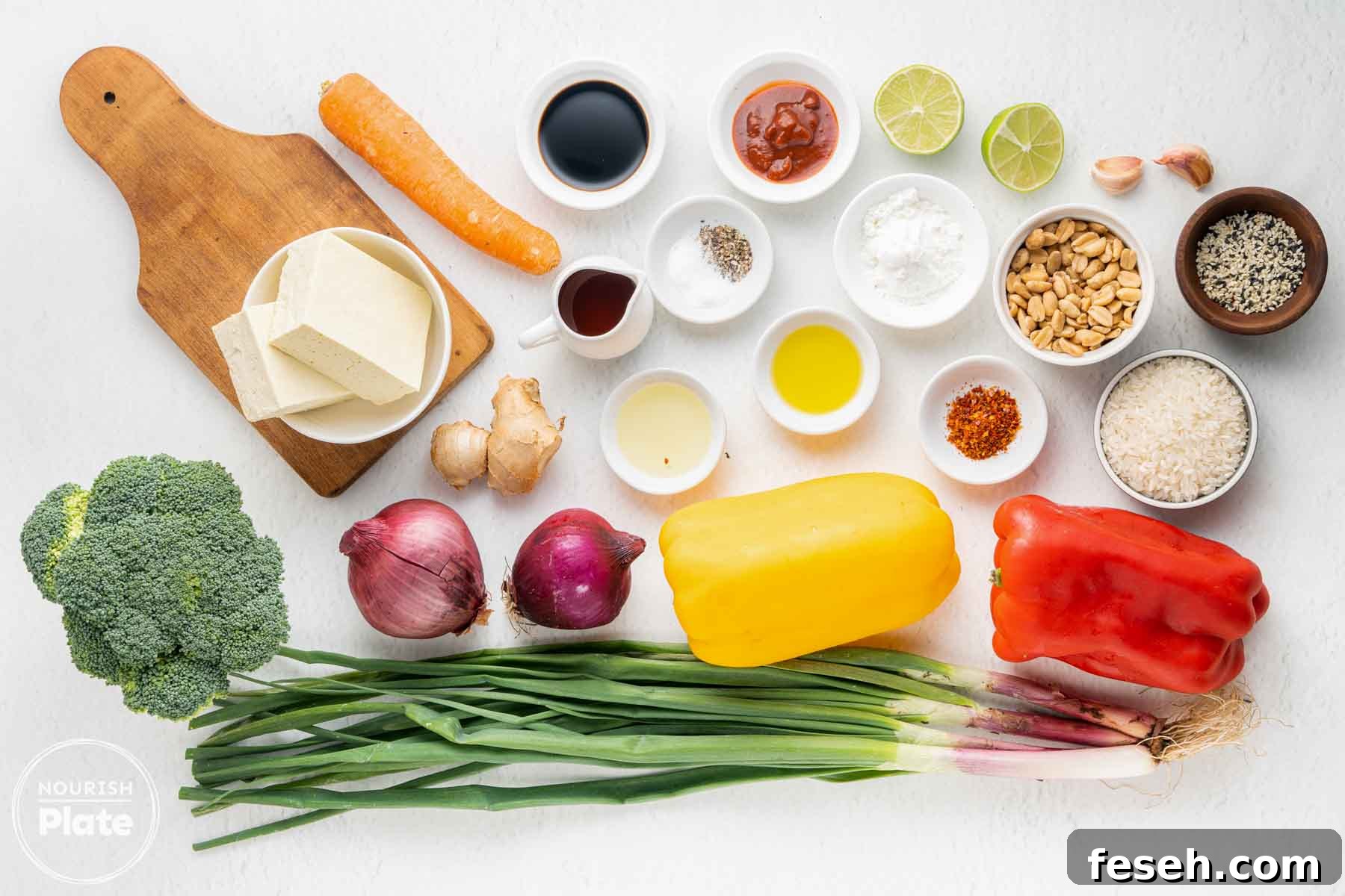 Overhead view of ingredients for crispy tofu stir fry including extra firm tofu, broccoli, carrot, red onion, green onions, red and yellow bell peppers, garlic, ginger, soy sauce, sriracha, maple syrup, olive oil, cornstarch, chili flakes, rice, sesame seeds, peanuts, and lime.