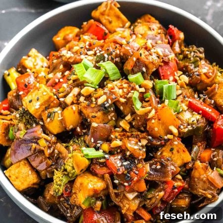 A bowl of crispy tofu stir fry with broccoli, red and yellow bell peppers, red onion, and carrot in a glossy dark sauce, topped with chopped peanuts, sesame seeds, and sliced green onions.