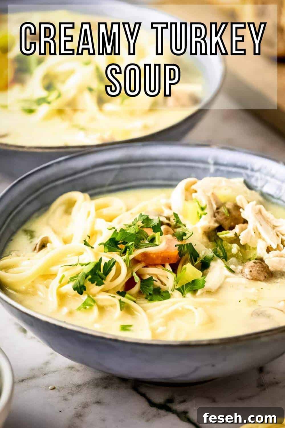 Side shot of two grey bowls with turkey noodle soup garnished with fresh herbs on a marble countertop with a wooden cutting board and sliced bread in the background.
