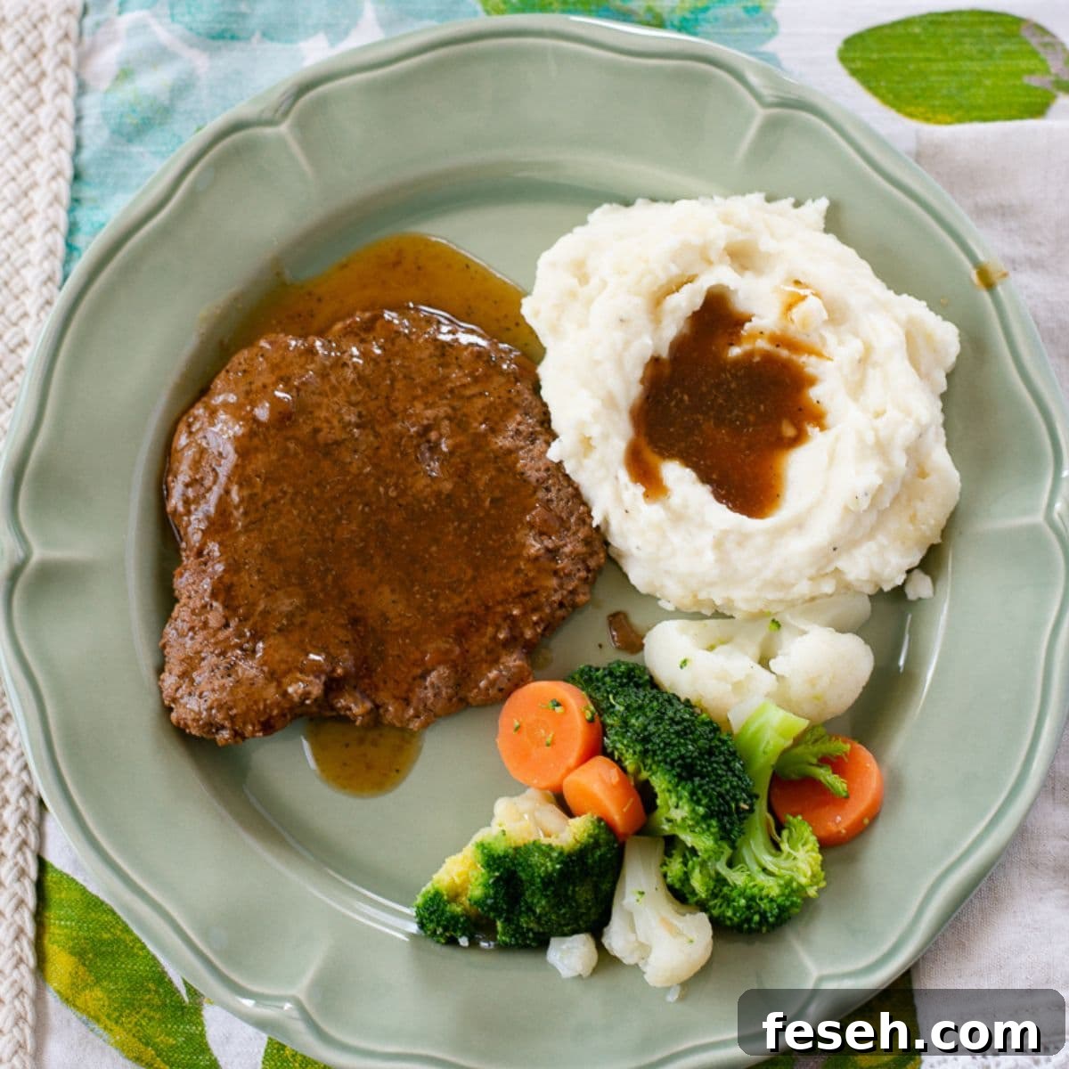A serving of Cube Steak smothered in gravy with mashed potatoes and vegetables on a dinner plate.