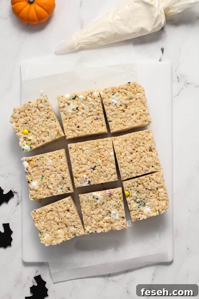 Unfrosted halloween rice krispie treats, sliced and set on a piece of parchment next to a bag of buttercream frosting