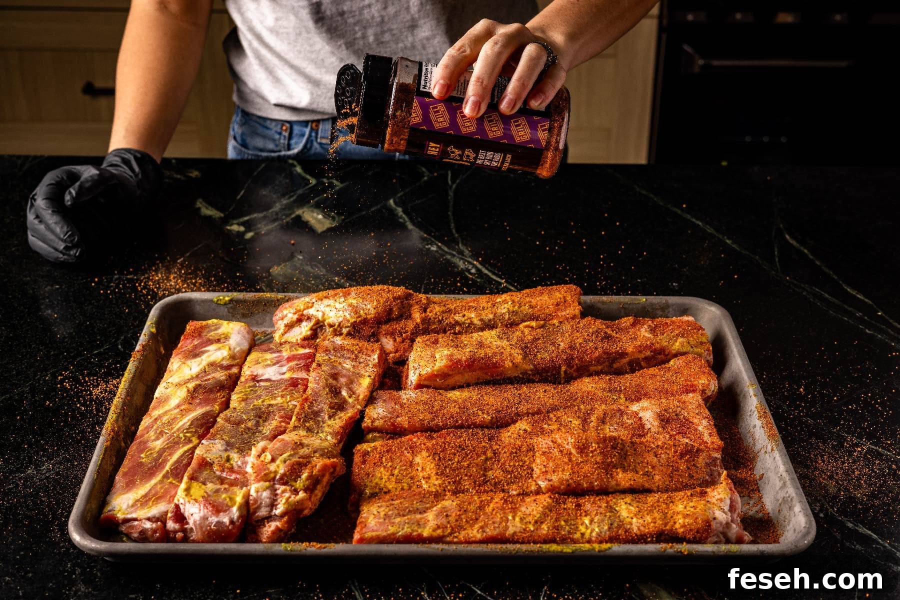 Uncooked ribs on baking sheet being sprinkled with seasoning.