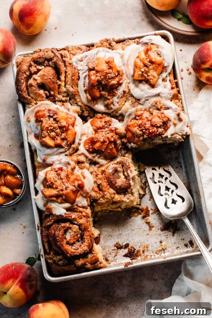 A wide angle photo of the cinnamon rolls in a pan, with some missing. Fresh peaches are scattered around the pan.
