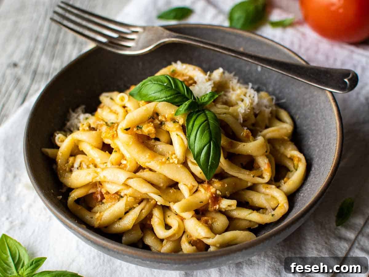 A grey bowl filled with handmade, twirled pasta with a reddish sauce, topped with a sprig of basil and a fork balance on the edge of the bowl.