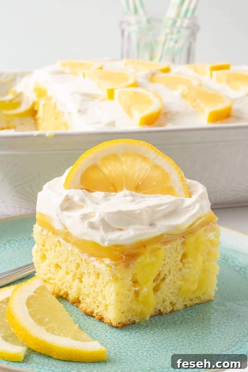 Lemon pudding poke cake on a plate with sliced lemons and remaining poke cake in the background.