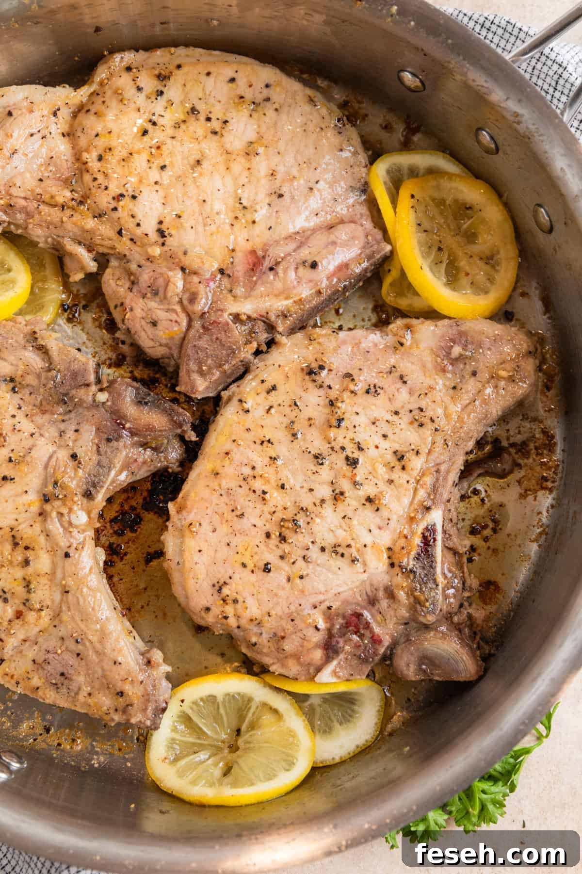 Lemon pepper pork chops cooking in stainless steel pan with lemon slices.