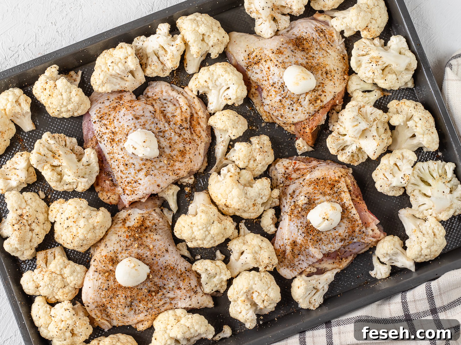 A baking sheet topped with a dollop of butter and cajun seasoned chicken thighs. Seasoned cauliflower surrounding the chicken and ready to go in the oven.