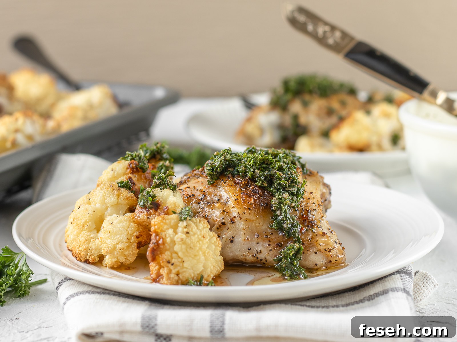 Table scene of baked chicken thighs and roasted cauliflower seasoned with a Cajun blend seasoning on plates and more on a baking sheet.