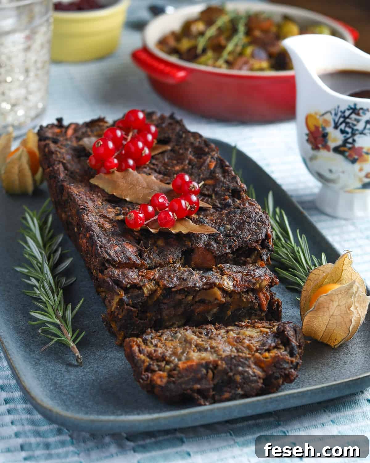 Photo of vegan & gluten-free nut roast on a blue-grey plate garnished with red currants, rosemary and cape gooseberries, on a blue tablecoth with a pretty jug of gravy to the side and a bowl of sprouts in the background.