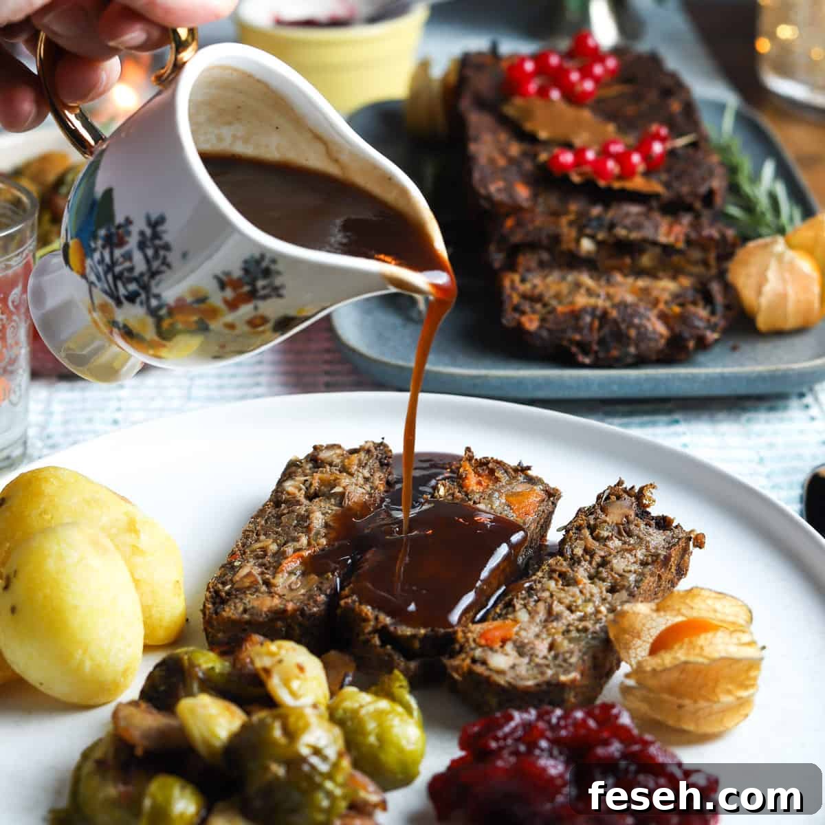 photo of a plate filled with 3 slices of this vegan nut roast, some roast potatoes, sprouts, cranberry sauce, and a pretty jug pouring gravy over the top, with the full vegan nut roast in the background.