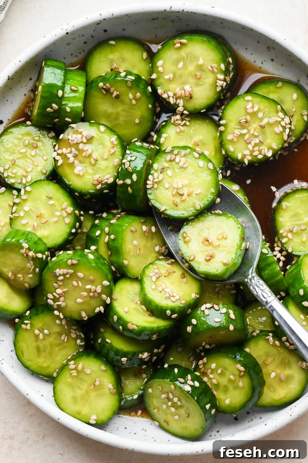 Prepared cucumber sesame salad in a shallow white speckled ceramic serving bowl, topped with sesame seeds, with a spoon lifting out several pieces of cucumber.
