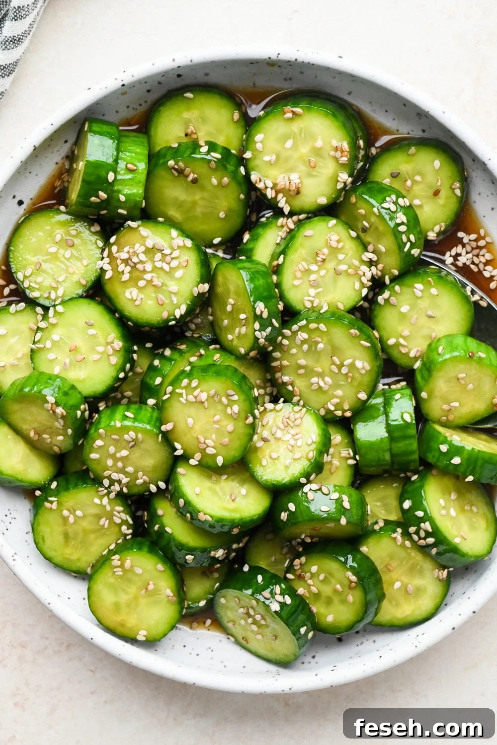 How to make cucumber sesame salad: Dressing poured over sliced cucumbers in a shallow ceramic serving bowl, after marinating and topping with additional sesame seeds.