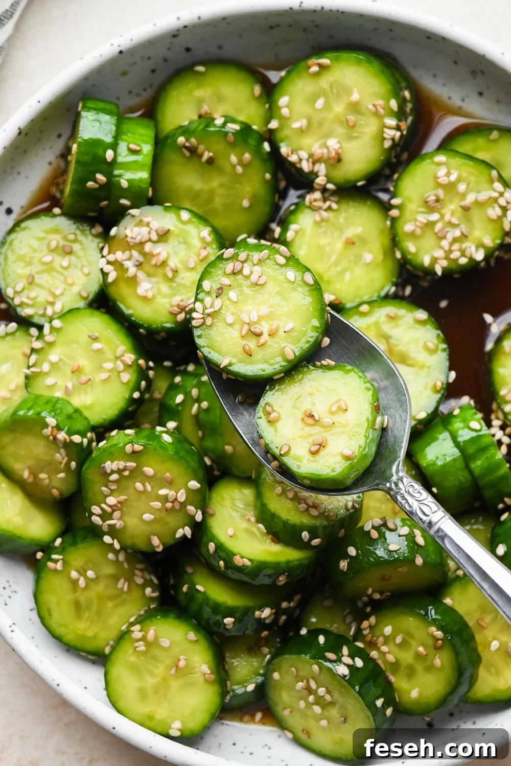 Prepared cucumber sesame salad in a shallow white speckled ceramic serving bowl, topped with sesame seeds, with a spoon lifting out several pieces of cucumber.