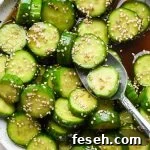 Prepared cucumber sesame salad in a shallow white speckled ceramic serving bowl, topped with sesame seeds, with a spoon lifting out several pieces of cucumber.