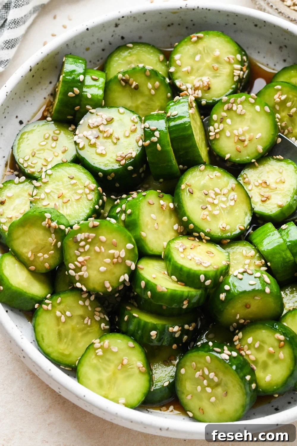Angled view of prepared cucumber sesame salad in a shallow white speckled ceramic serving bowl, topped with sesame seeds.