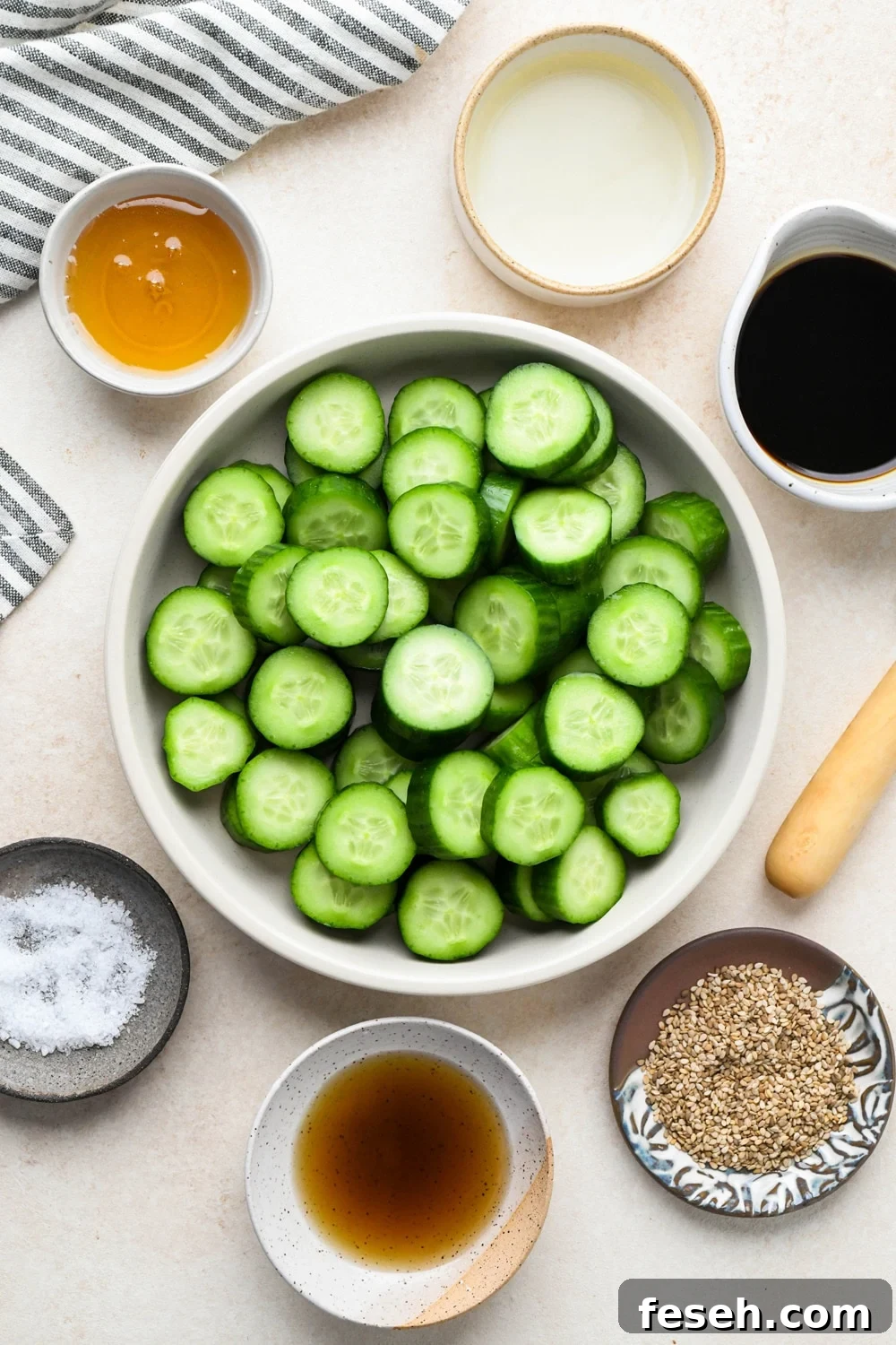 Ingredients for cucumber sesame salad in various ceramic dishes on a light cream background, with a striped blue and white linen.