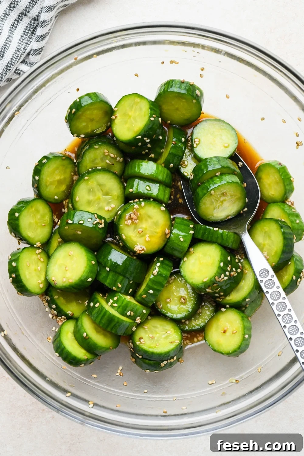 How to make cucumber sesame salad: Dressing poured over sliced cucumbers in a medium sized glass bowl, after marinating.