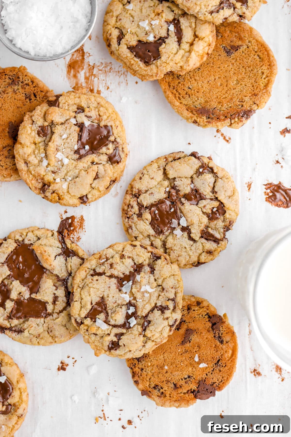 cropped overhead shot of eight chocolate chip cookies on chocolate stained parchment paper with glass of milk and measuring cup of salt around.