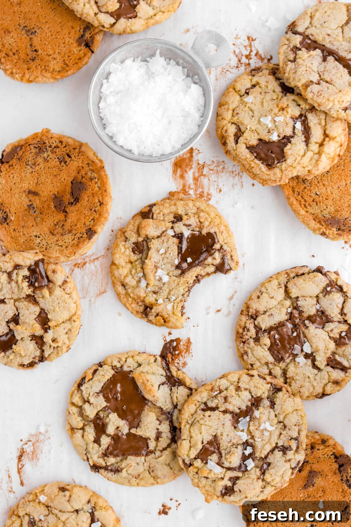 overhead shot of chocolate chip cookies on chocolate stained parchment paper with measuring cup of salt above cookie with bite missing.