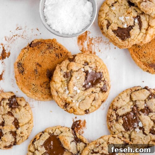 close up overhead shot of chocolate chip cookies on parchment paper with flaked salt on top of four cookies with measuring cup of flaked salt above.