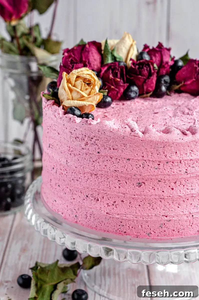 A pink frosted blueberry layer cake. It has blueberry speckles in the buttercream frosting and the cake is topped with beige and dark red/pink roses and fresh dark blue blueberries.