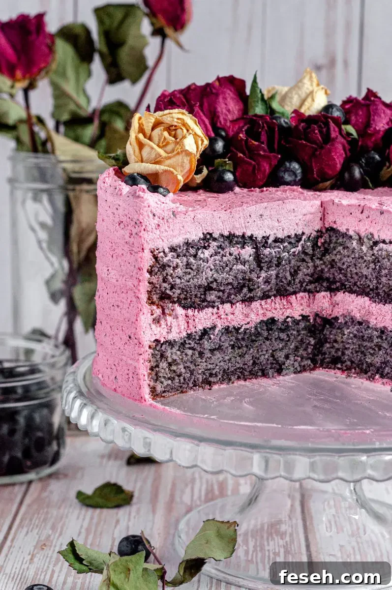 A pink frosted blueberry layer cake. It has blueberry speckles in the buttercream frosting and the cake is topped with beige and dark red/pink roses and fresh dark blue blueberries. There are several slices cut out of the cake revealing the inside.