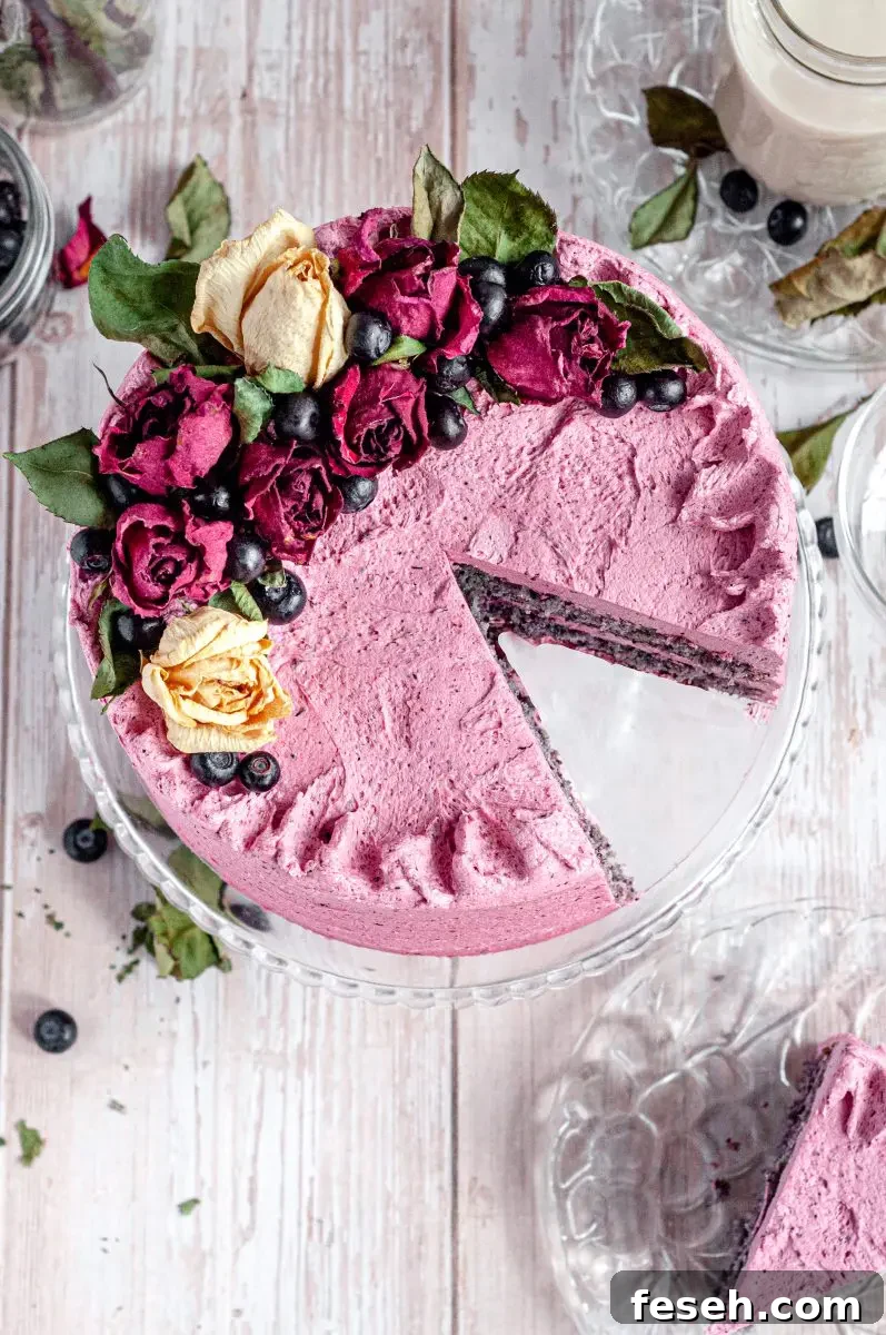 The top view of a frosted blueberry layer cake. The blueberry frosting is a pink color with speckles of blueberries in it. The cake is topped with dried beige and red roses and dark blue blueberries. There is a slice cut out of the cake and it is sitting on a plate beside the cake.