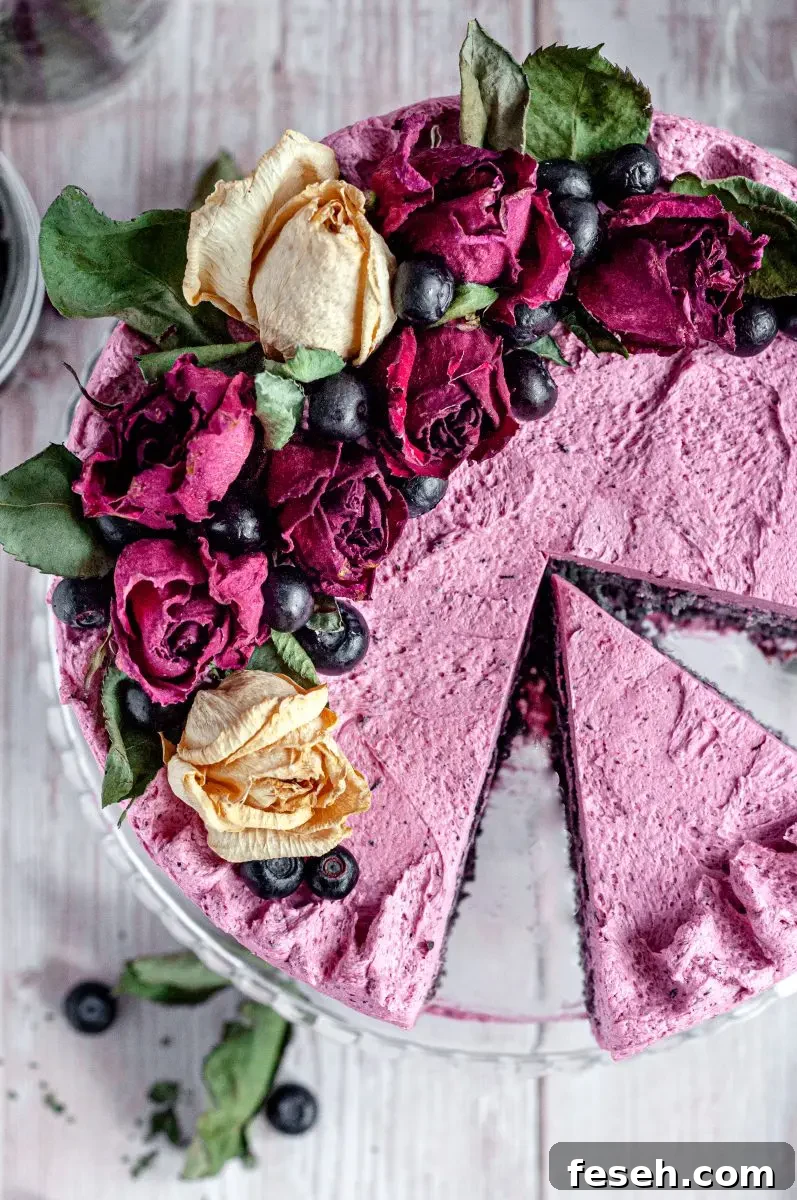 The top view of a frosted blueberry layer cake. The blueberry frosting is a pink color with speckles of blueberries in it. The cake is topped with dried beige and red roses and dark blue blueberries. There are two slices cut out of the cake and a slice of cake is sitting in between where the two slices have been cut out.