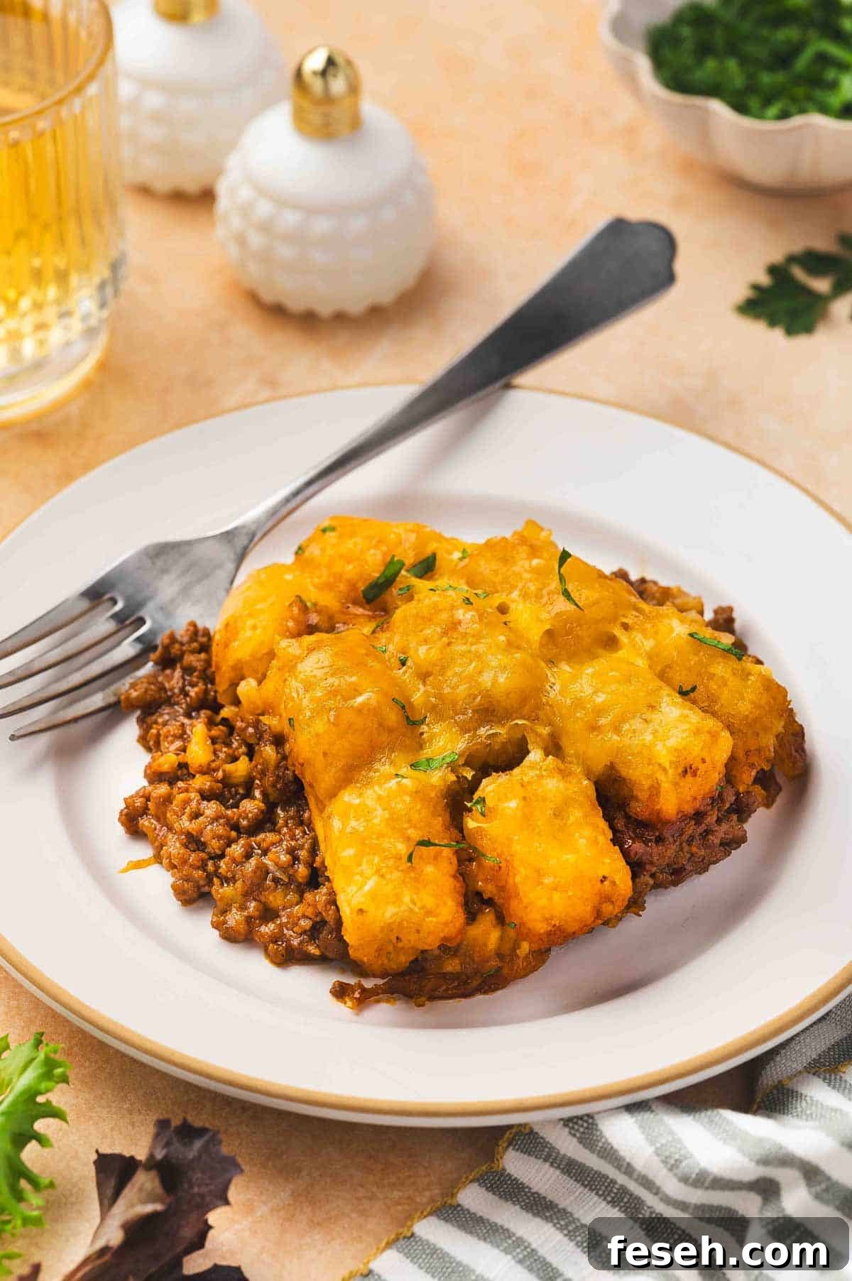A plate with a serving of sloppy joe tater tot casserole with a fork with chopped parsley garnish.