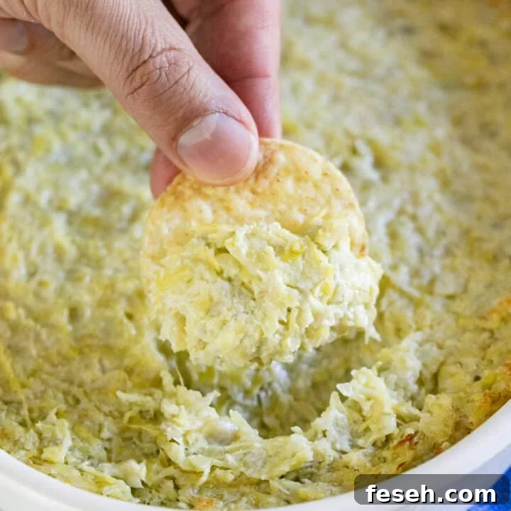A hand pulling a chip with artichoke dip out of a pan of it.