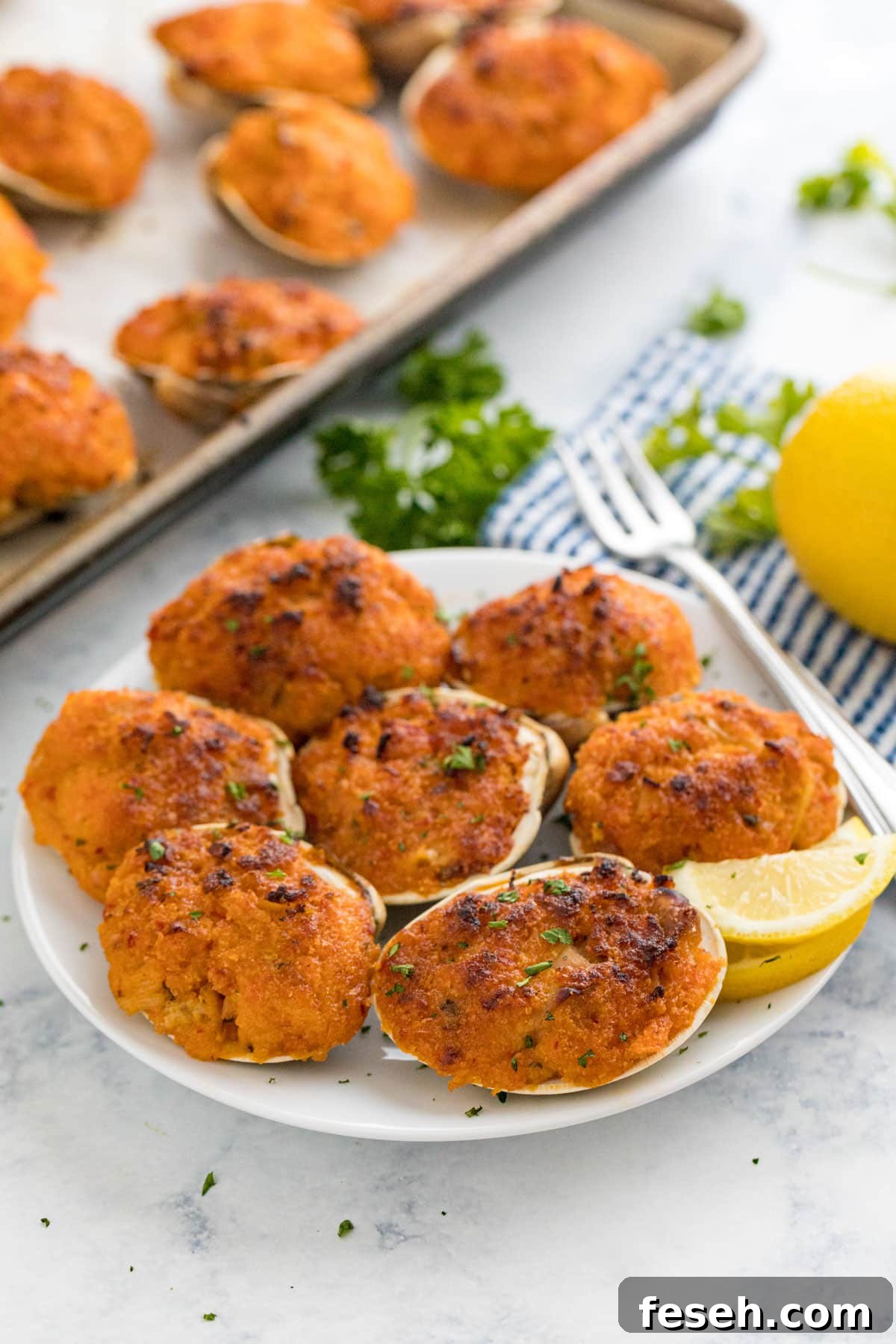 A plate of baked clams with a cocktail fork and lemon wedge on it with the pan, a striped napkin, lemon and parsley behind it.